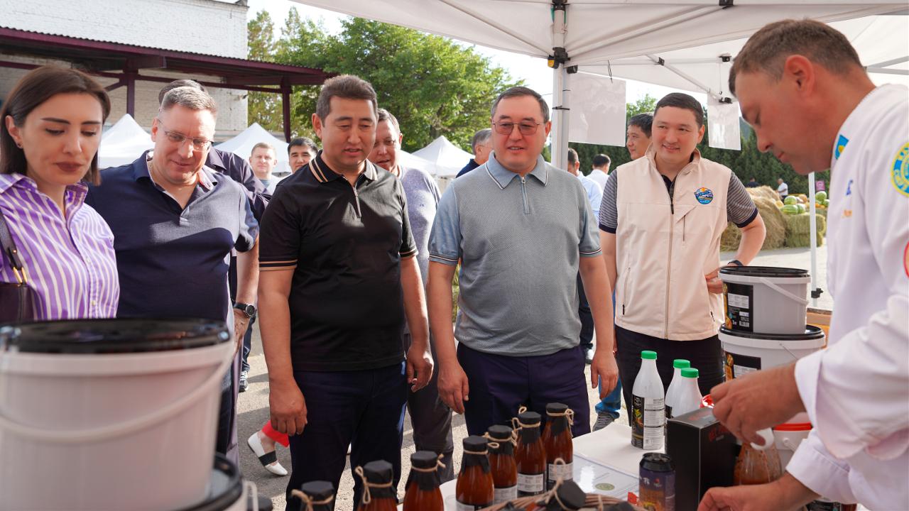Group of people at an outdoor market stall, examining bottled sauces/beverages laid out on a table under a tent.