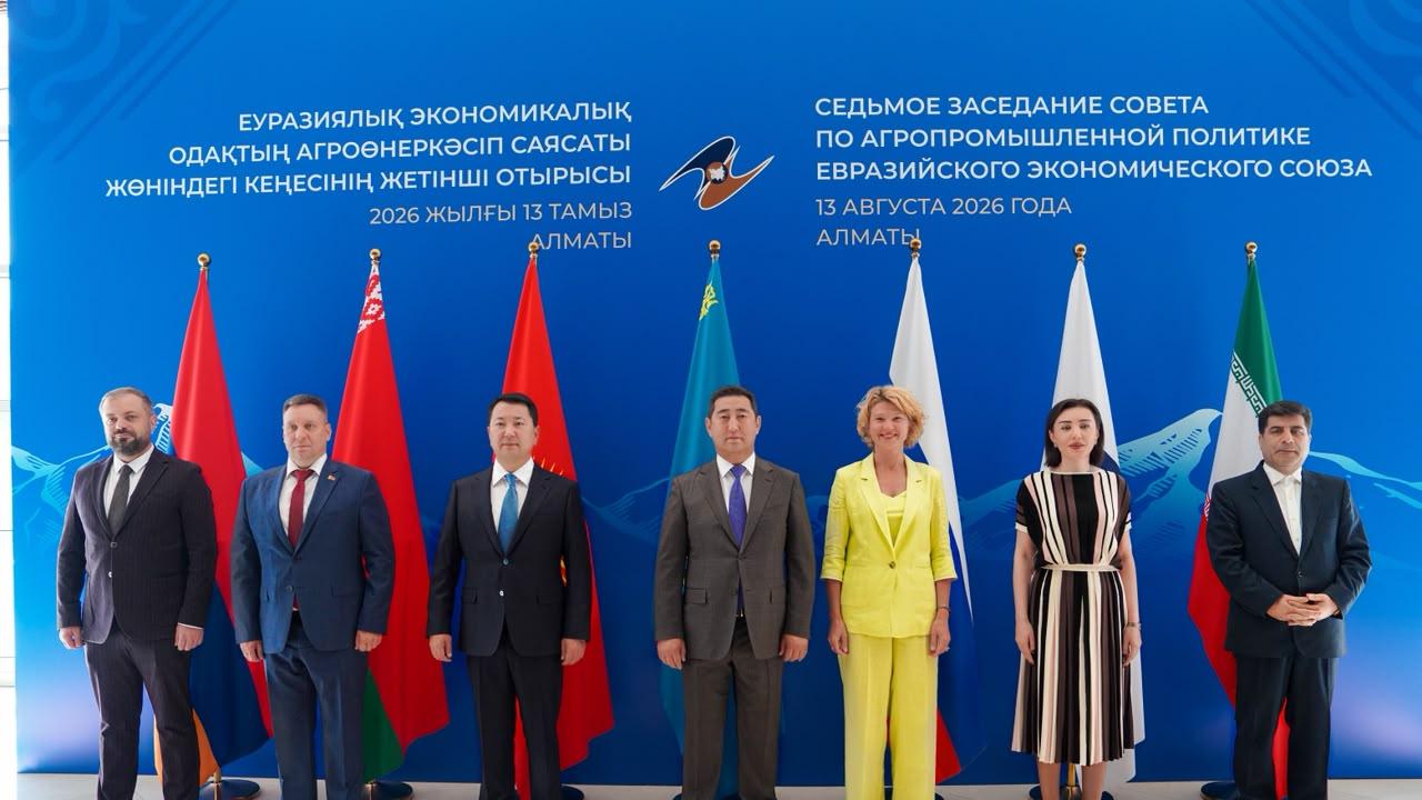Group of seven officials in business attire standing in front of a blue backdrop with flags, during an international economic meeting.